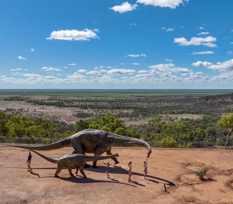 Children and adults standing next to large dinosaur statues on a plateau overlooking the Winton outback.