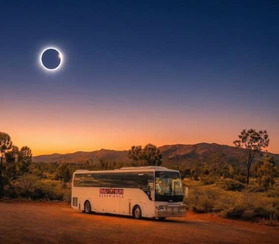 A white Emu Run Experience tour coach parked on a red dirt road in the Alice Springs outback during a dramatic total solar eclipse. Credit: Emu Run Experience