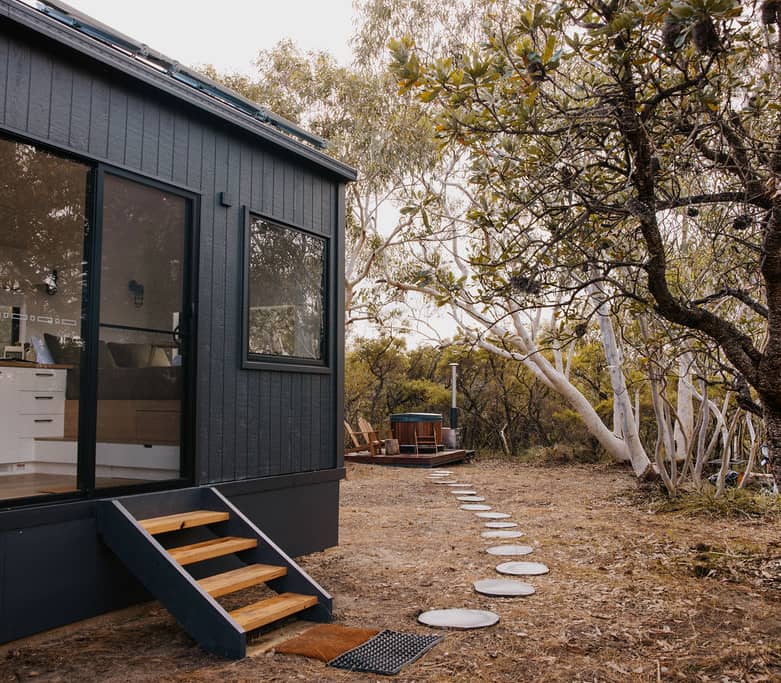 Interior view of a tiny home showing a clean white kitchen and a bed by a large window.