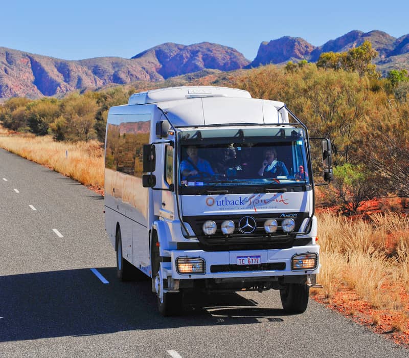 A white Mercedes-Benz all-terrain touring coach for Outback Spirit Tours drives on a paved road through a dry, mountainous outback landscape under a clear blue sky. The coach has a license plate readin