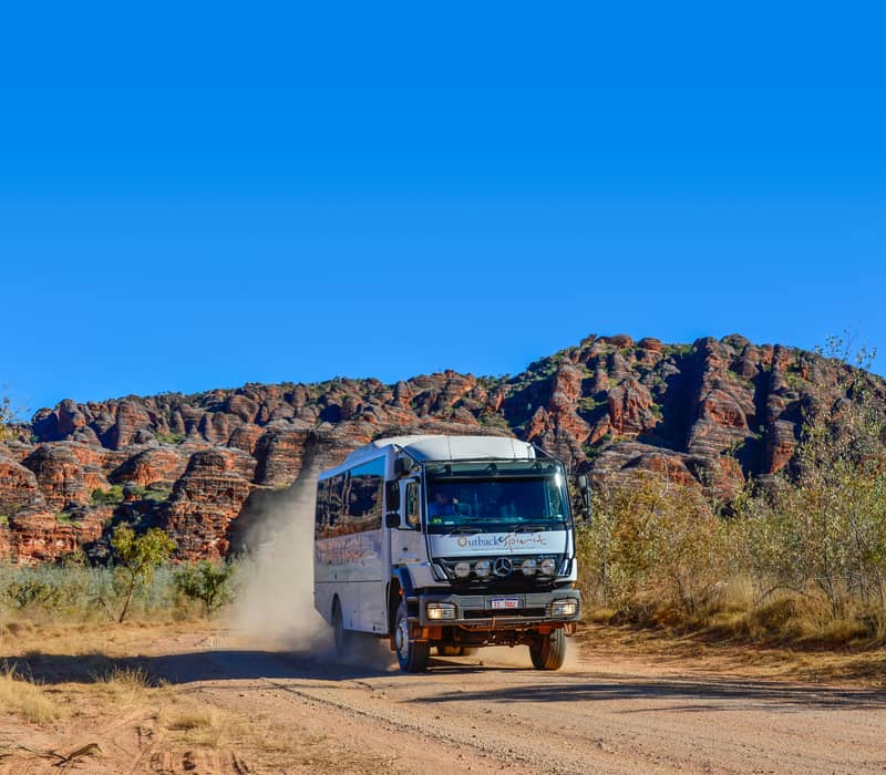 A white and tan Outback Spirit safari coach driving along a dusty track with the iconic orange and black striped beehive domes of the Bungle Bungle Range in the background.
