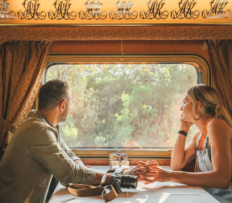 Interior view of a Gold Service Lounge on a Journey Beyond rail journey. A man and woman sitting at a white-clothed dining table inside a luxury Journey Beyond train carriage, looking out the window at green bushland.
