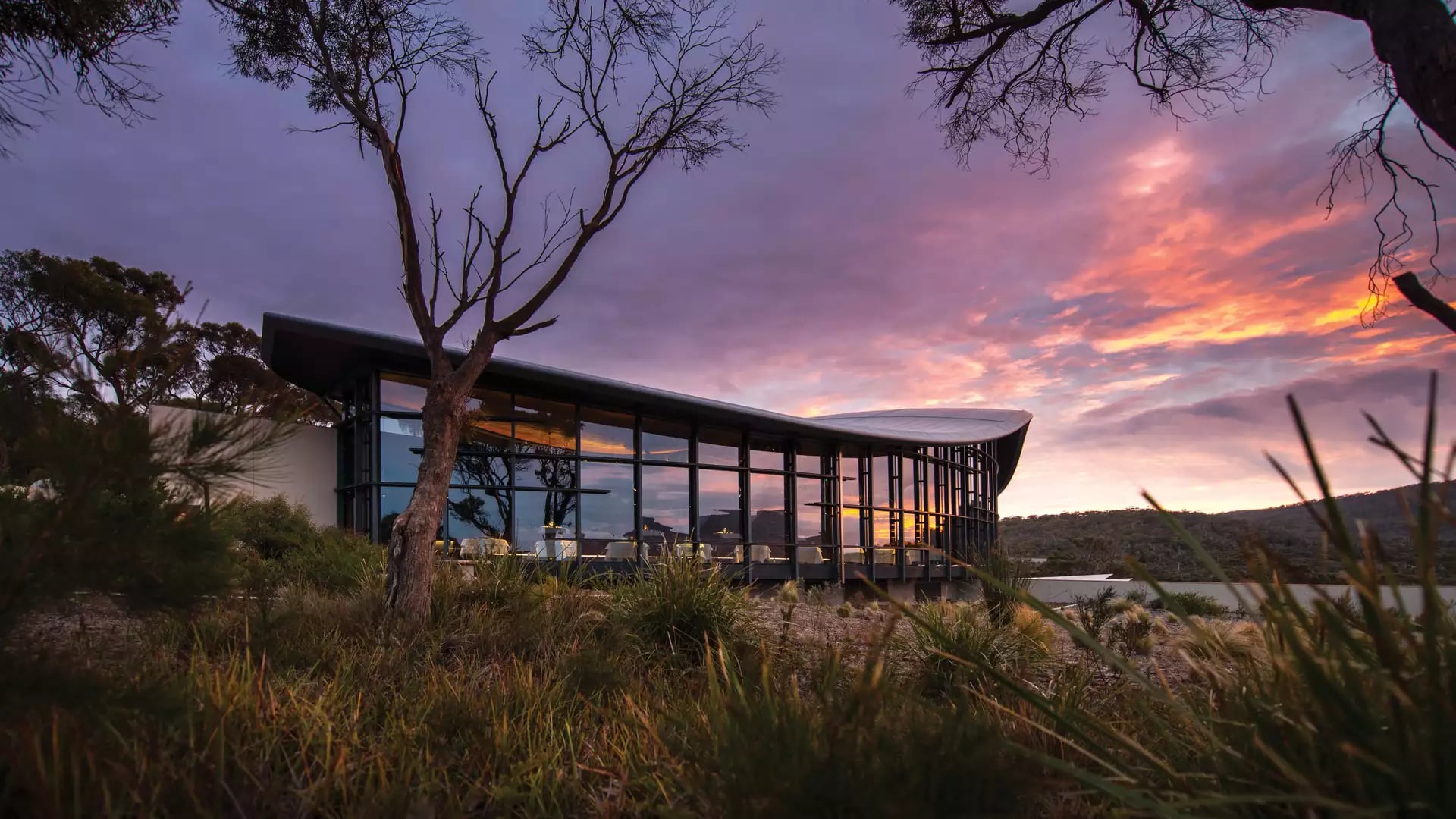An external evening view of the Palate Restaurant at Saffire Freycinet, showcasing the iconic curved roof and floor-to-ceiling glass windows illuminated against a vibrant Tasmanian sunset.
