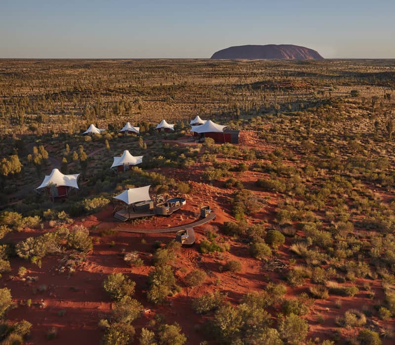 High-angle shot of sixteen white peaked-roof tents arranged in a curve along bright orange sand dunes, with sparse green desert oaks and the large red rock of Uluru on the horizon.