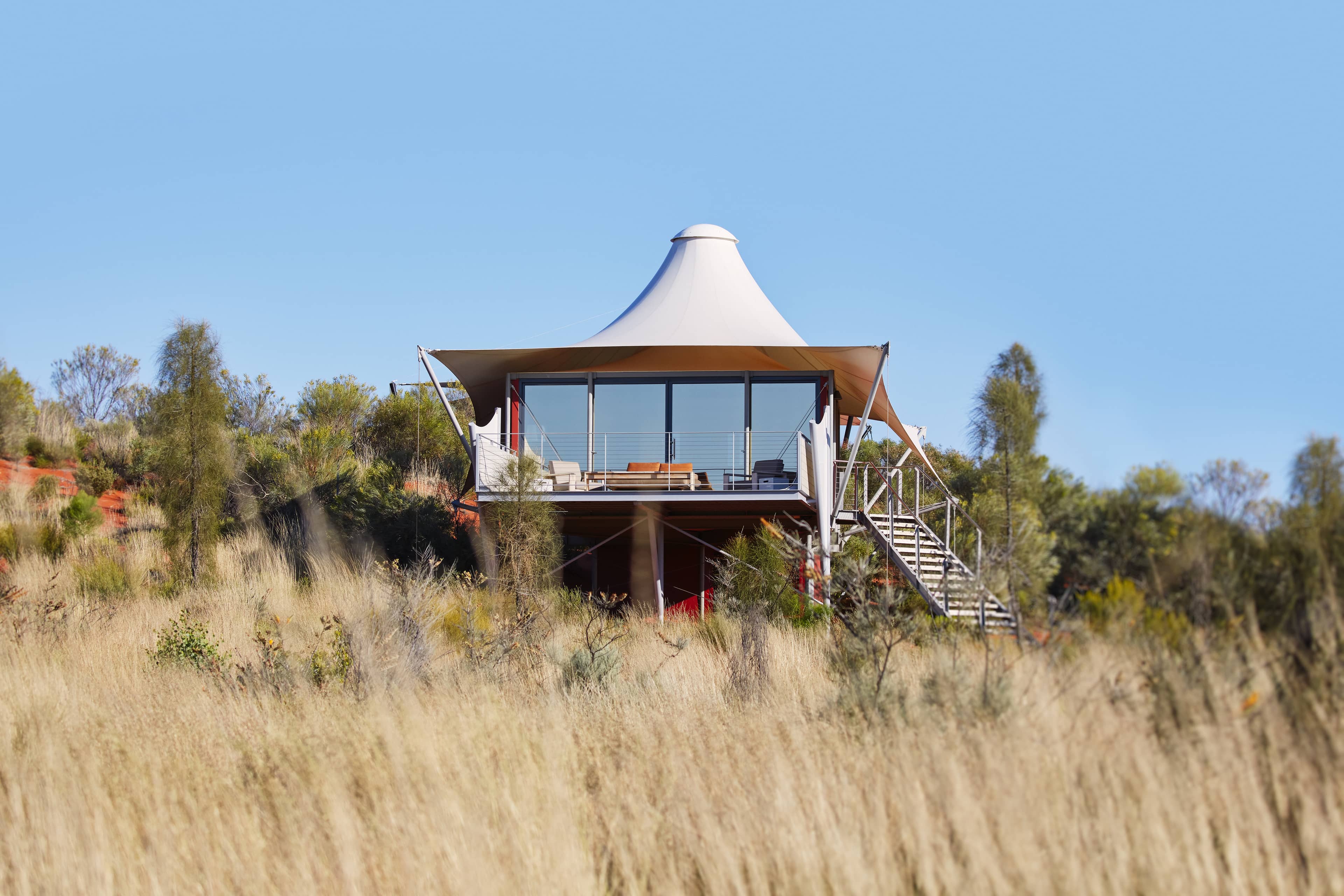 A low-angle exterior view of a luxury elevated tent at Longitude 131°, featuring a distinctive white peaked canopy and floor-to-ceiling glass walls that provide panoramic views of the desert.
