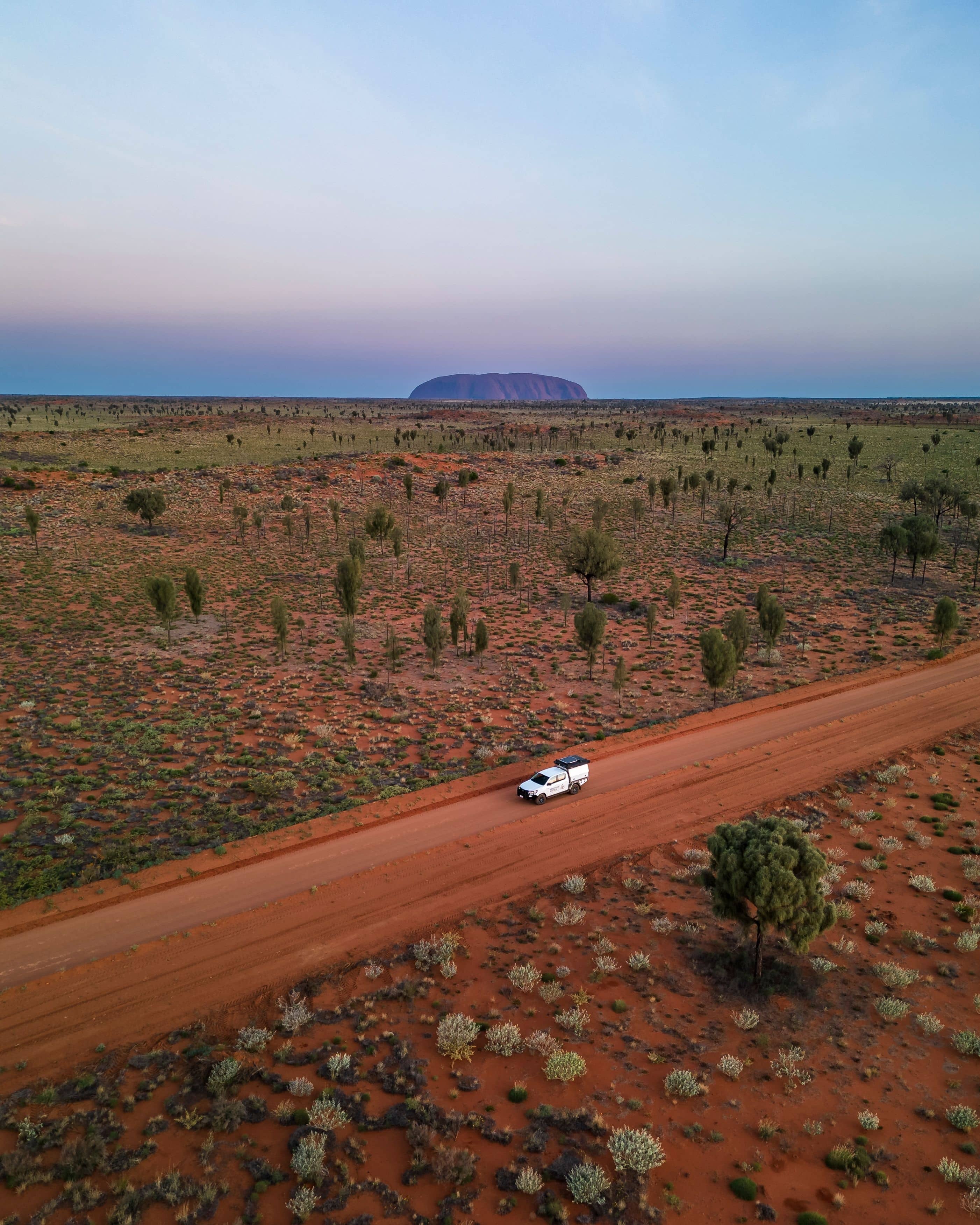 A white 4WD vehicle travels along a remote red dirt track through the vast desert landscape of Uluru-Kata Tjuta National Park, with the iconic silhouette of Uluru visible on the horizon.