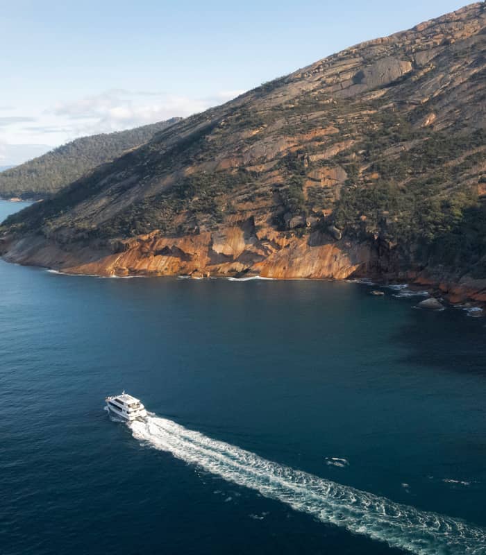 Aerial view of a white tour boat leaving a white wake as it travels past steep orange granite cliffs.