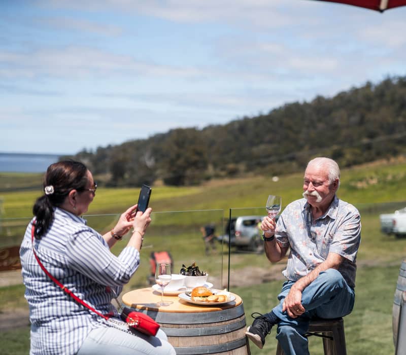 A man with a white mustache enjoying a glass of white wine at a barrel table outdoors at Devil's Corner, while a woman takes his photo.