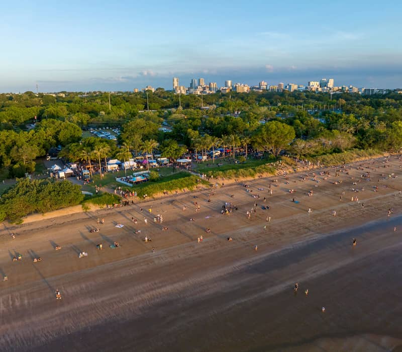 A high-angle aerial view of the Mindil Beach Sunset Markets, showing the bustling stalls nestled in the trees and crowds gathered on the sand toward the Darwin city skyline. Credit: Tourism & Events NT/Tourism Australia