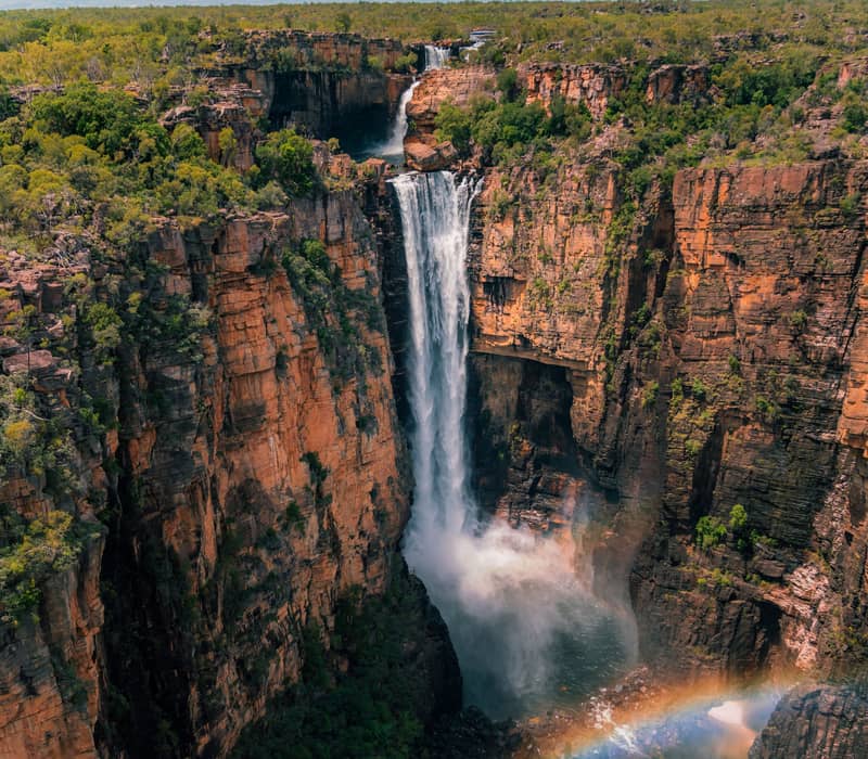 A spectacular aerial view of Jim Jim Falls during the tropical summer, showing the massive waterfall plunging over the Arnhem Land escarpment with a rainbow appearing in the mist.