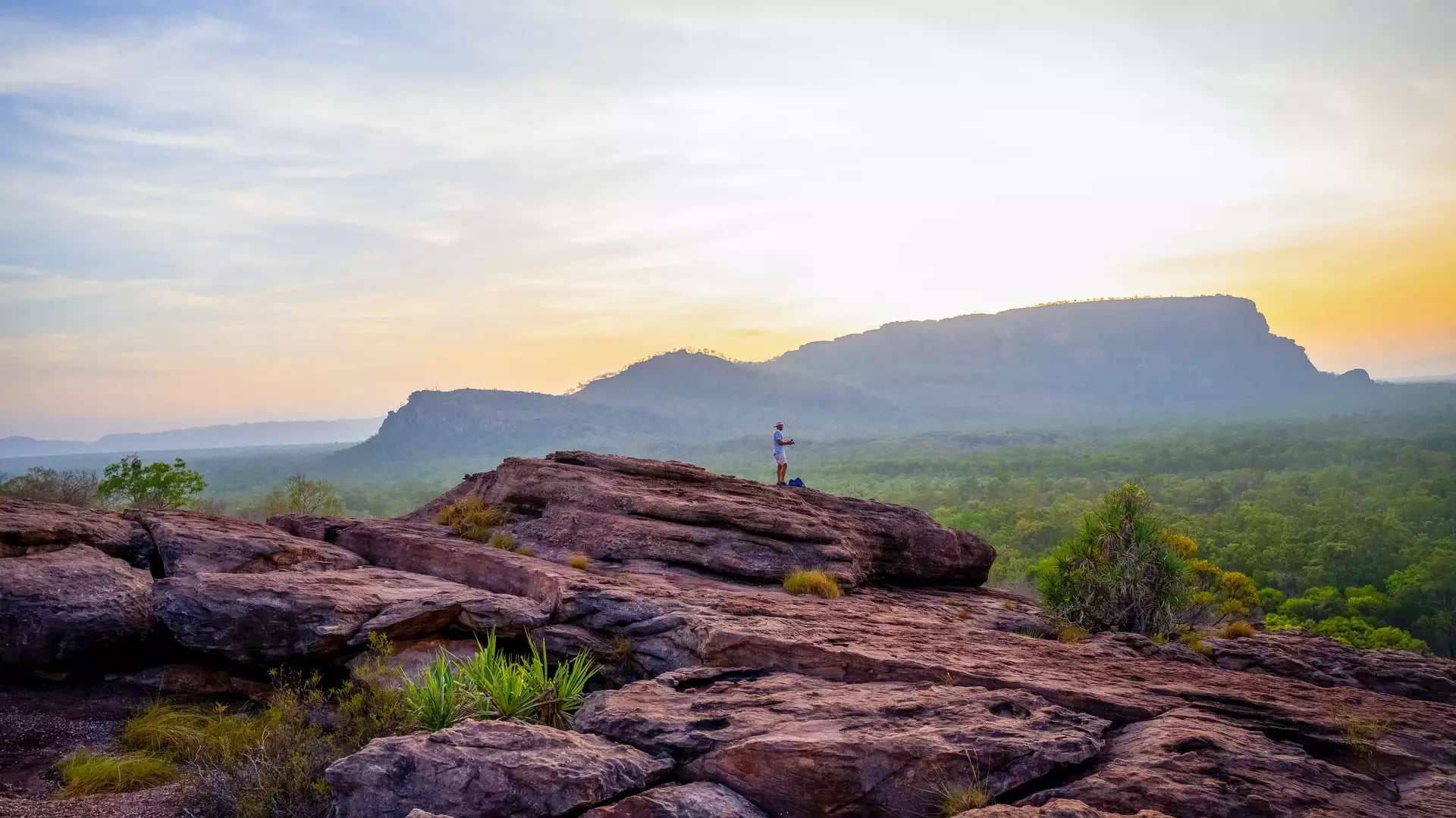 A photographer stands atop the rugged ironstone slopes of Nawurlandja Lookout, capturing the sunset glow over Anbangbang Billabong and the distant Nourlangie rock massif.
