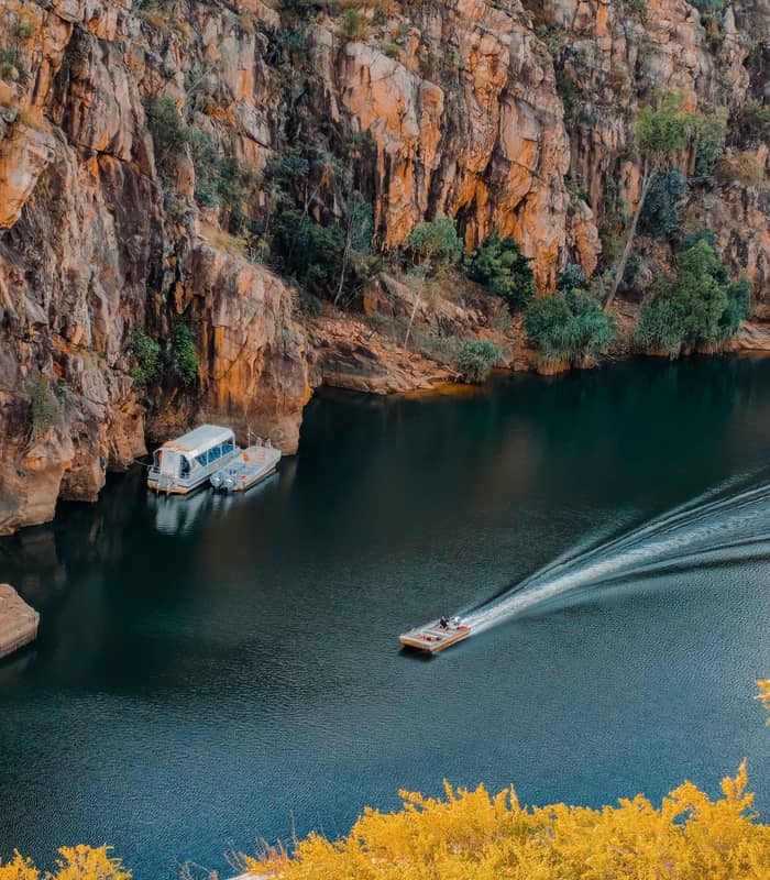 A vertical photograph taken from a high vantage point overlooking a wide river. The river flows through a deep valley with steep, rocky orange cliffs on one side and a dense green forest on the other. The sky above is a pale, hazy blue, and the sunlight highlights the rugged textures of the stone walls.