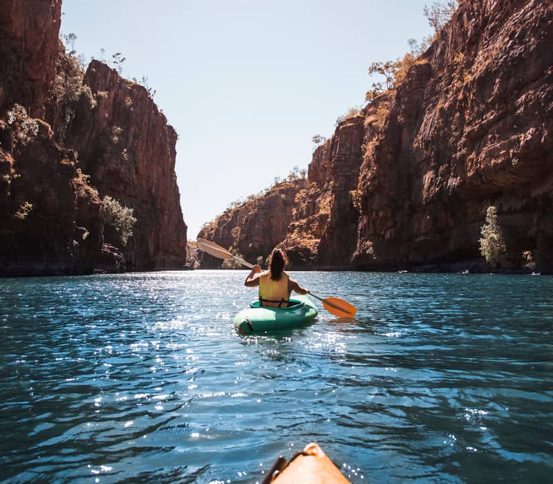 A first-person perspective of a kayaker paddling through the calm, blue waters of Nitmiluk Gorge, framed by sheer sandstone cliffs under a bright midday sun.