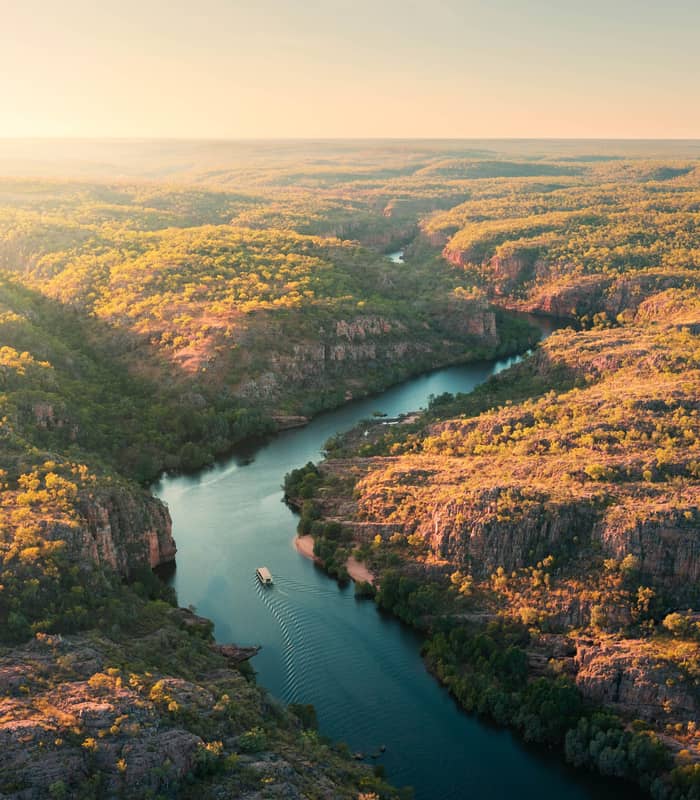 A flat-bottomed tour boat navigates the calm, green-tinted waters of Nitmiluk (Katherine) Gorge, dwarfed by the massive, weathered sandstone escarpments that rise vertically from the river's edge.