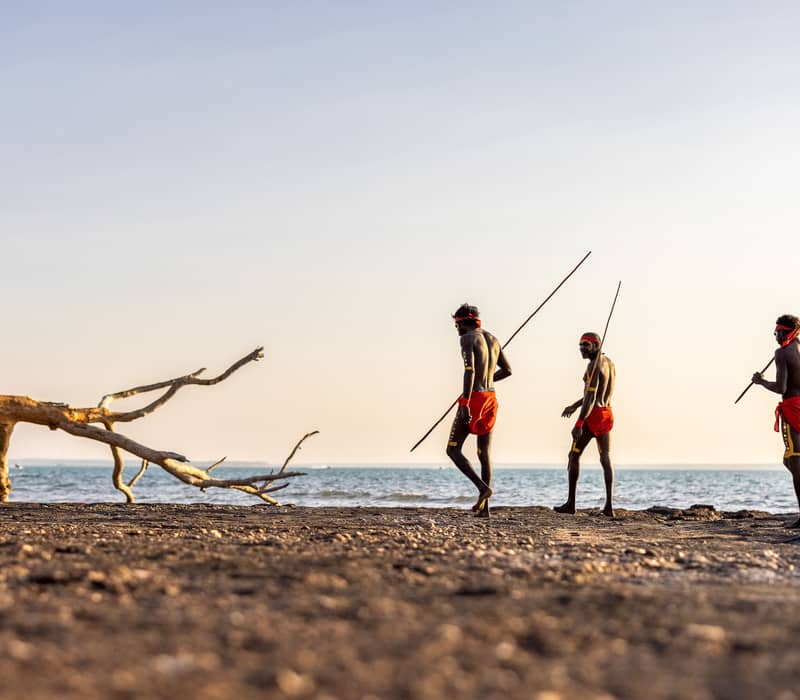 Larrakia men in traditional ceremonial attire walk along the Darwin shoreline, carrying spears against a soft morning light.