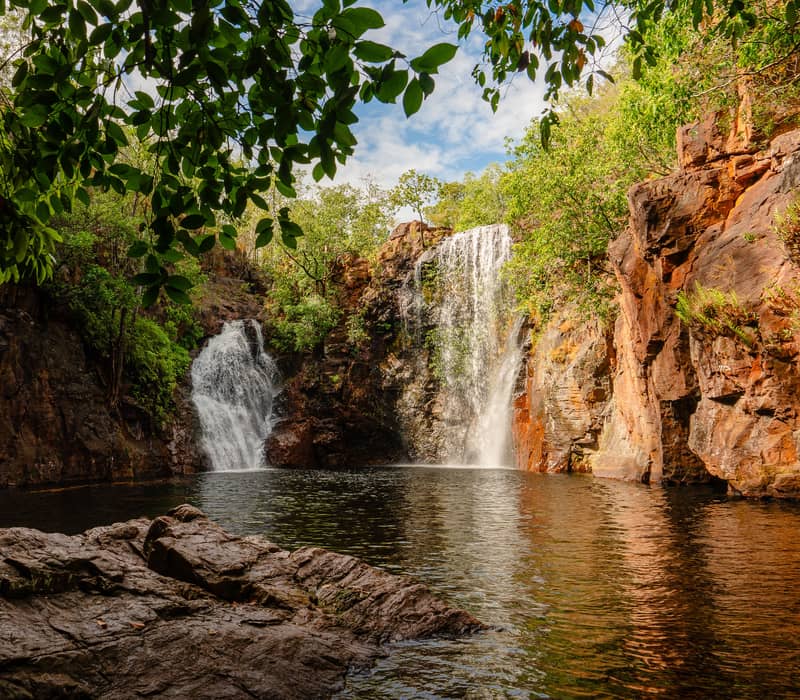 A scenic view of the twin cascades of Florence Falls, framed by lush overhanging foliage and rugged red sandstone cliffs in Litchfield National Park.