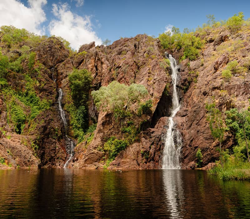 A wide panoramic view of Wangi Falls, featuring two large cascades flowing over a massive rock face into a wide, accessible swimming pool.