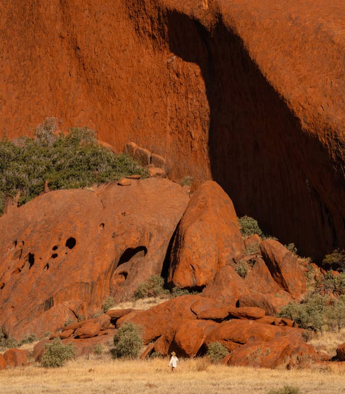 Alone hiker is dwarfed by the sheer scale of the towering red sandstone walls and unique geological textures of Uluru's base.