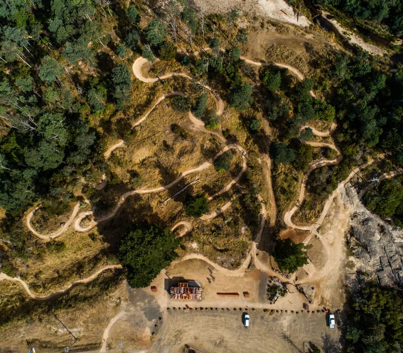 A high-altitude overhead shot shows a series of winding, light-brown dirt mountain bike trails snaking through a landscape of dense green forest and patches of dry, golden-brown scrubland. A small clearing with parked vehicles and a small building with a