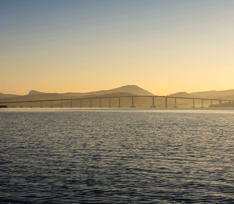 Aerial view of a long bridge connecting coastal city suburbs divided by a large body of water at dusk.