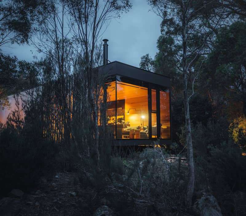 A modern, rectangular cabin (The Retreat) with floor-to-ceiling glass walls is nestled deep within a dark forest at twilight. The interior of the cabin glows with a warm, golden light, revealing a cozy living space, while the surrounding trees and shrubs are silhouetted against a moody, dark blue sky.