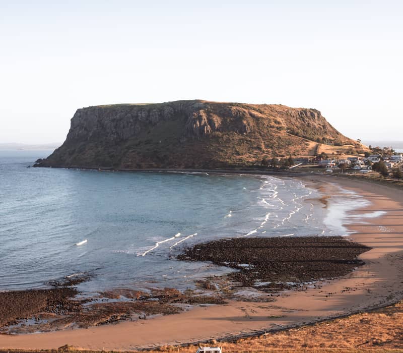 Massive flat-topped volcanic rock formation overlooking a crescent beach and a yellow van on a coastal road.