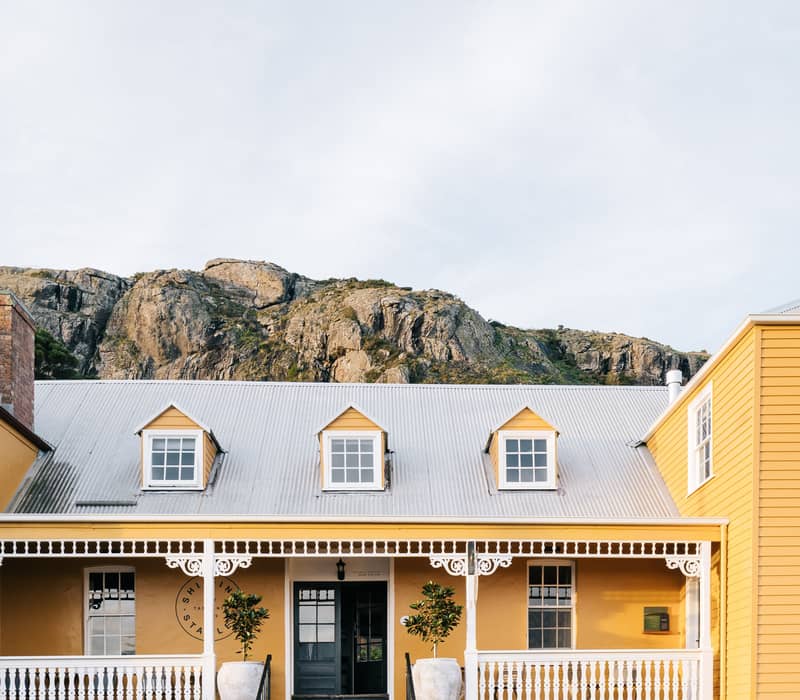 A charming two-story yellow heritage building with a silver corrugated iron roof and three small dormer windows. It features a long white veranda with decorative wooden trim and sits behind a low stone wall. In the background, the massive, sheer rocky cliff face of "The Nut" rises high above the town under a clear sky.
