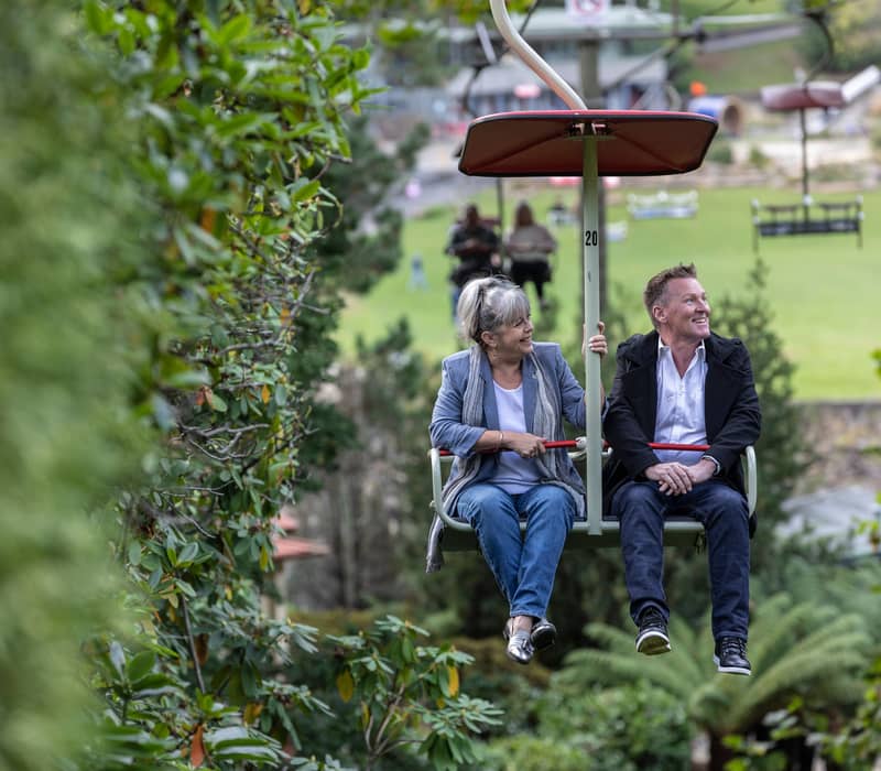 A smiling couple riding a green and red chairlift, passing closely by lush green trees with the reserve grounds visible in the background.