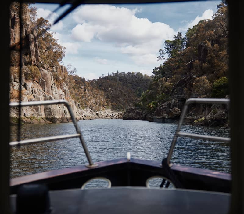 View from the front of a boat looking down a narrow river canyon with high, rocky cliffs and green trees under a bright sky.