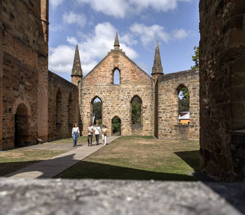 Four people walking on a wooden boardwalk inside the roofless shell of a large stone church with pointed windows and high spires.