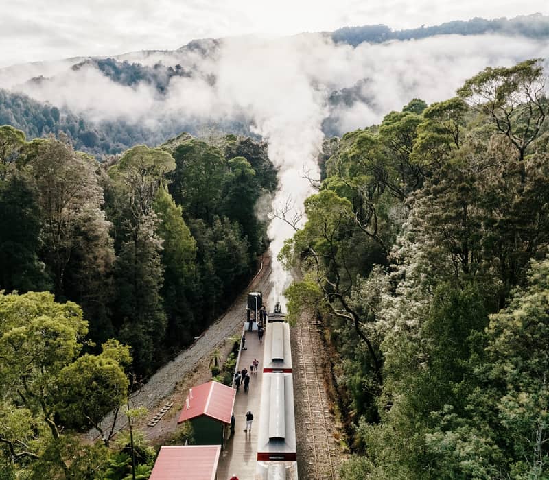 High-angle view of a steam train at a forest station surrounded by mist-covered green mountains.