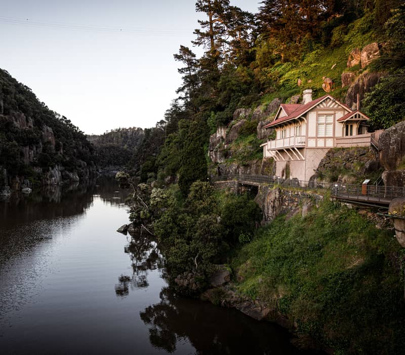 A high-angle view looking down the South Esk River at Cataract Gorge, featuring the historic white and red-roofed Cataract Cottage perched on a steep, lush green hillside overlooking the calm dark water