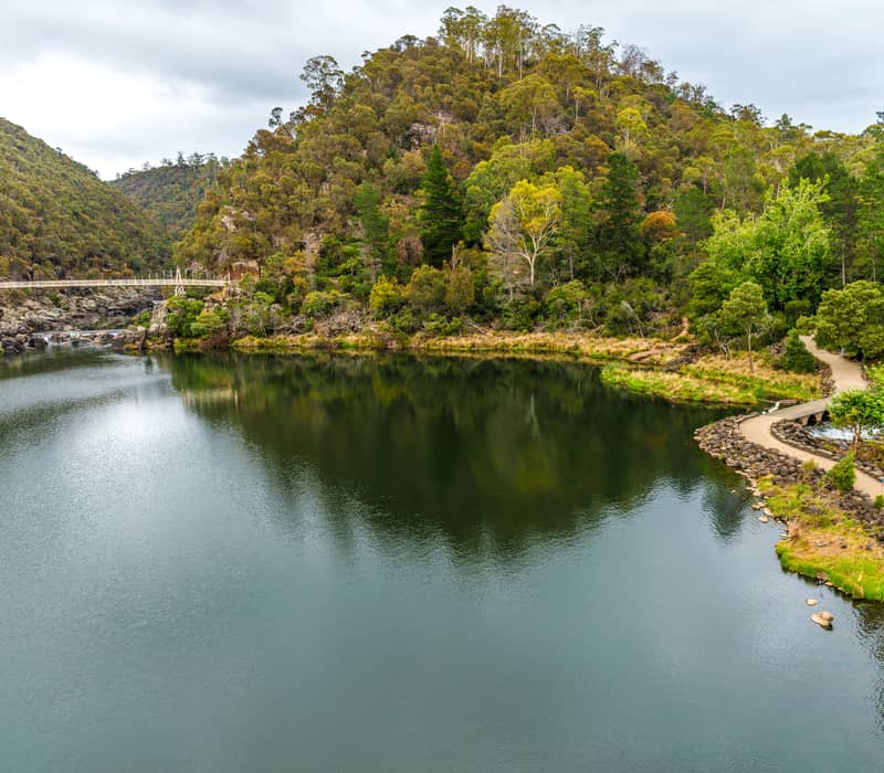 Wide landscape view of the Alexandra Suspension Bridge at Cataract Gorge, reflected perfectly in the still, misty waters of the South Esk River.
