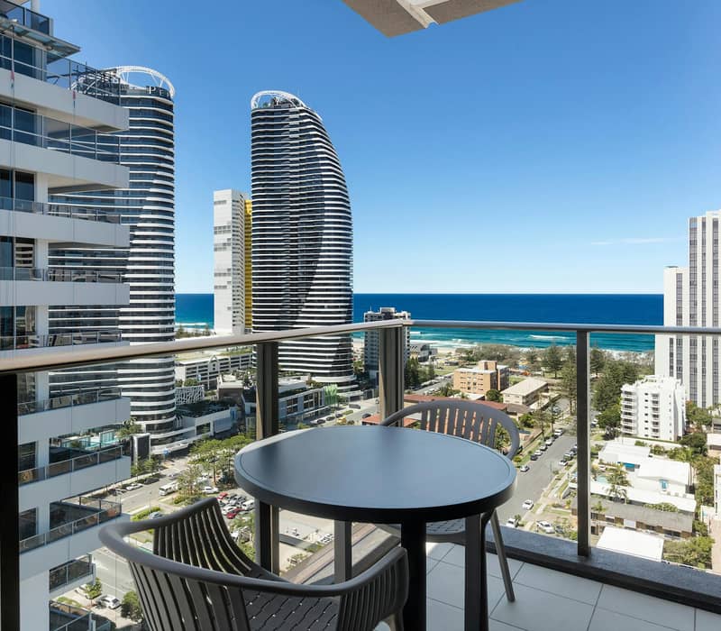 Hotel bedroom with large windows showing ocean and city view, king bed with pink runner and pillow.