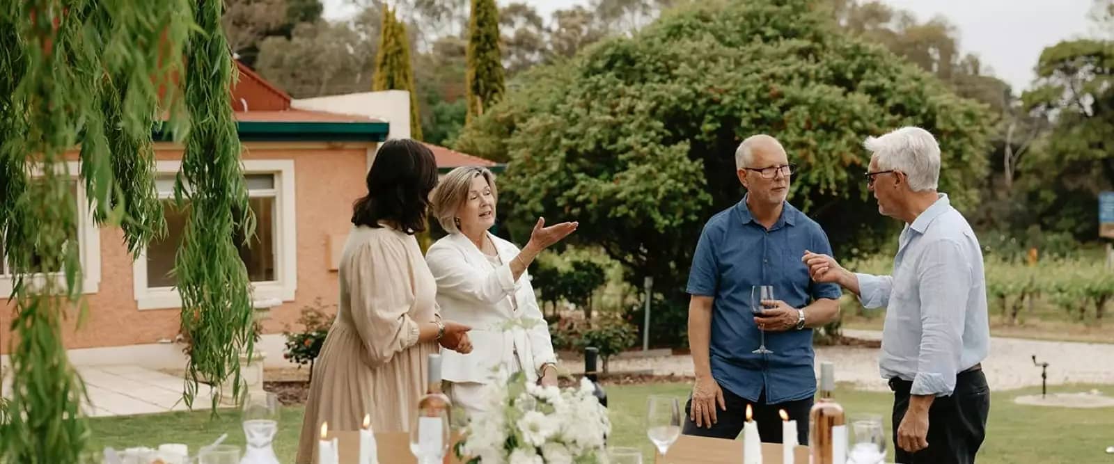Four people socializing outdoors near a table set with wine and candles in a garden party setting.