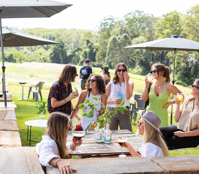 Friends drinking and chatting outdoors under umbrellas at a rustic wooden table.