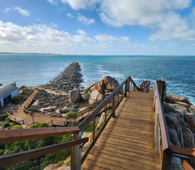 Wooden boardwalk leading down to rocky coastline with ocean and blue sky in the background.