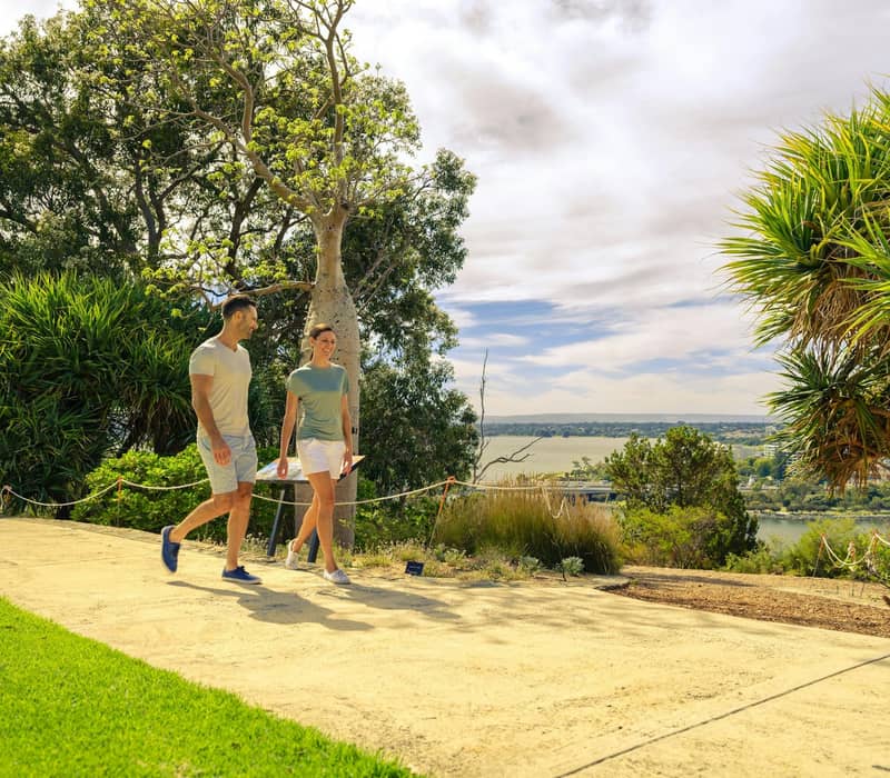 Couple walking on a path surrounded by trees and greenery with a scenic view in the background.