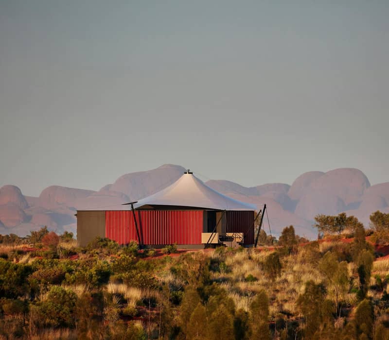 Luxury desert tent with white roof and red walls in Australian outback with distant rock formations.