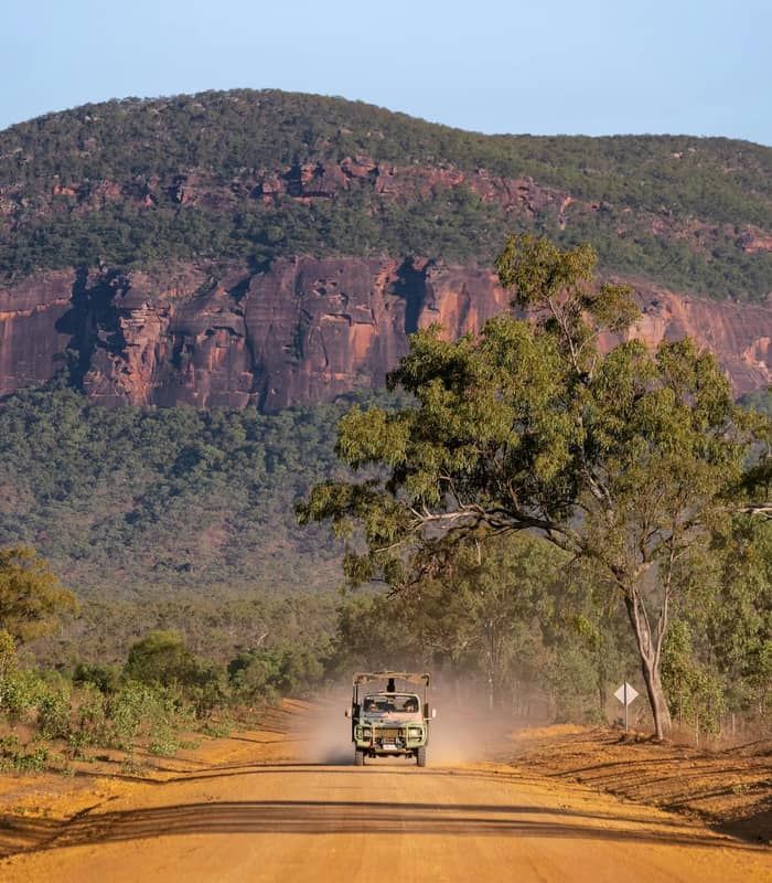 Truck on dusty dirt road with mountain and trees in background under clear sky