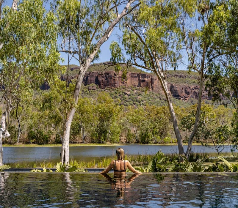 Woman in pool looking at river, trees, and rocky mountain under blue sky.