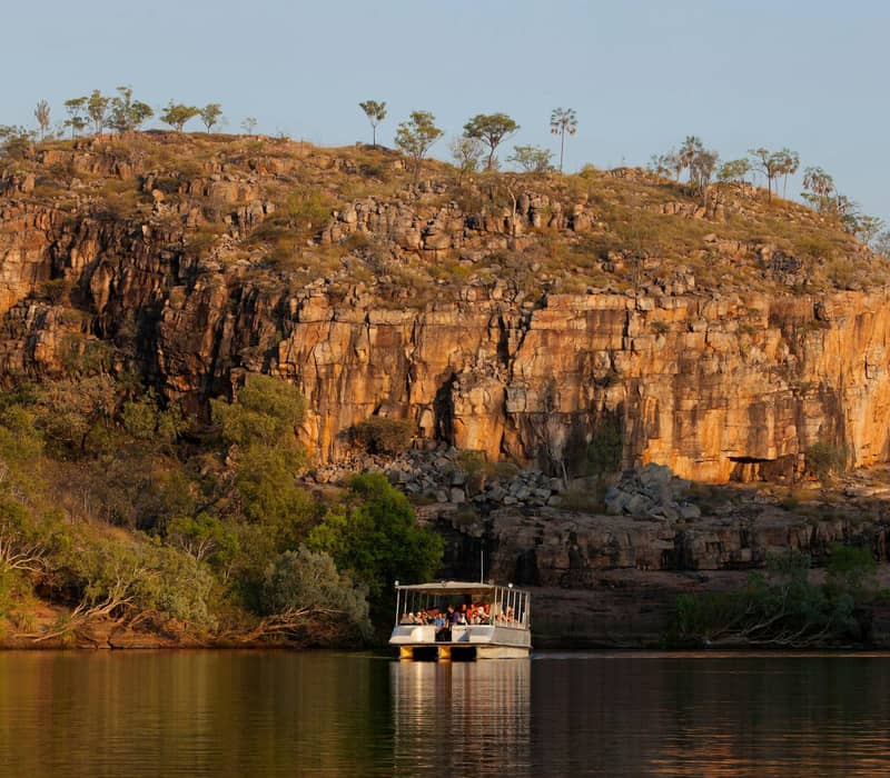 Tour boat cruising on river near rocky cliffs and trees at sunset in Nitmiluk Gorge