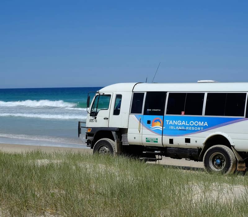 White 4WD bus parked on sandy beach with ocean waves and clear blue sky.