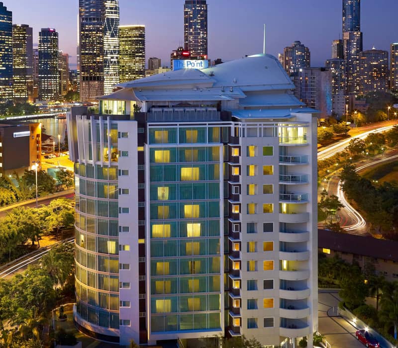 The Point Brisbane Hotel lit up at night with tall city buildings in the background and light trails from traffic.