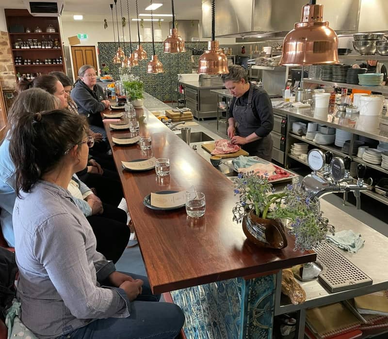 People seated at a counter watching a chef prepare meat in a modern kitchen.