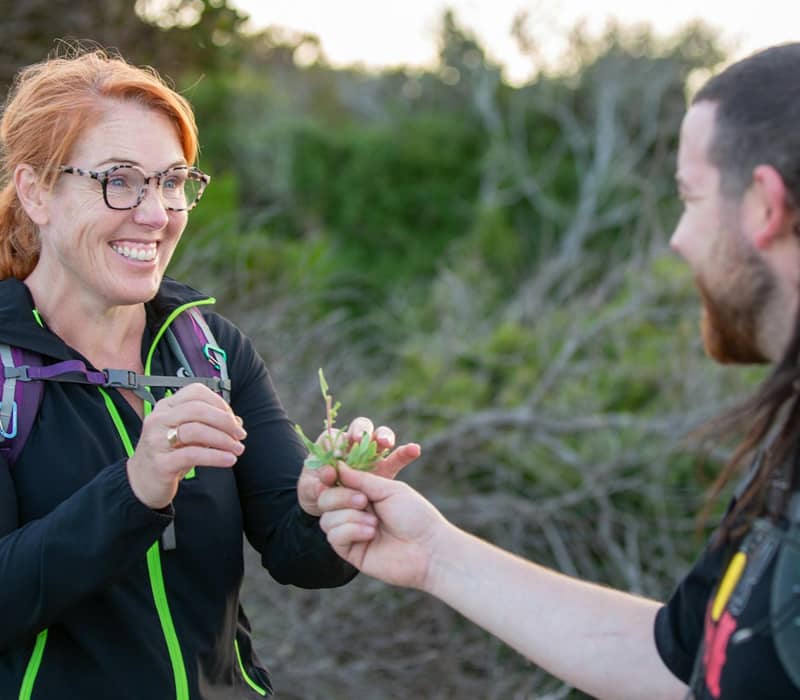Two hikers smiling and sharing a plant during a walk in nature.